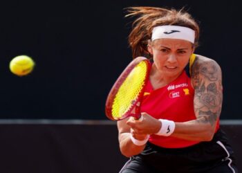 Tenista española golpeando la pelota durante un partido, con camiseta roja de la selección y fondo oscuro.