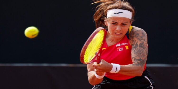 Tenista española golpeando la pelota durante un partido, con camiseta roja de la selección y fondo oscuro.